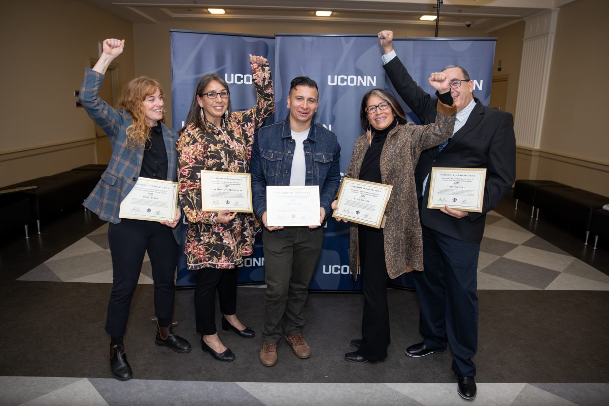 Five people posing for a picture in front of a UConn sign with their hands up.