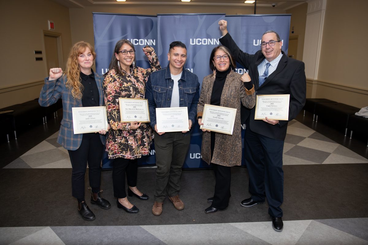 Five people posing for a picture in front of a UConn sign.