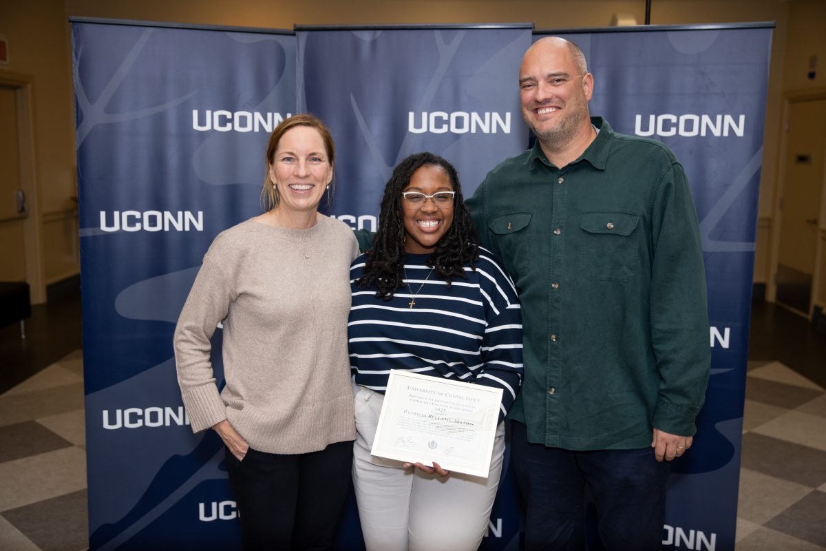Three people posing for a picture in front of a Uconn sign.