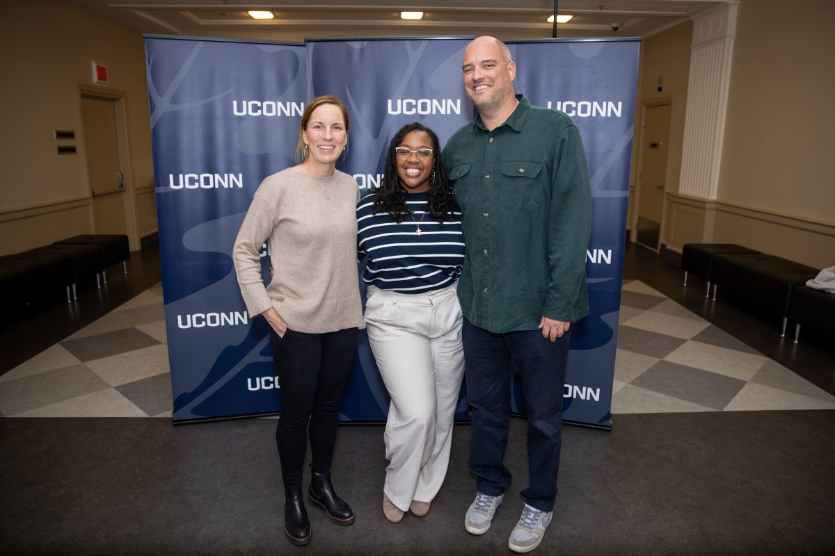 Two ladies and a gentleman posing for a picture with a UConn sign behind them.