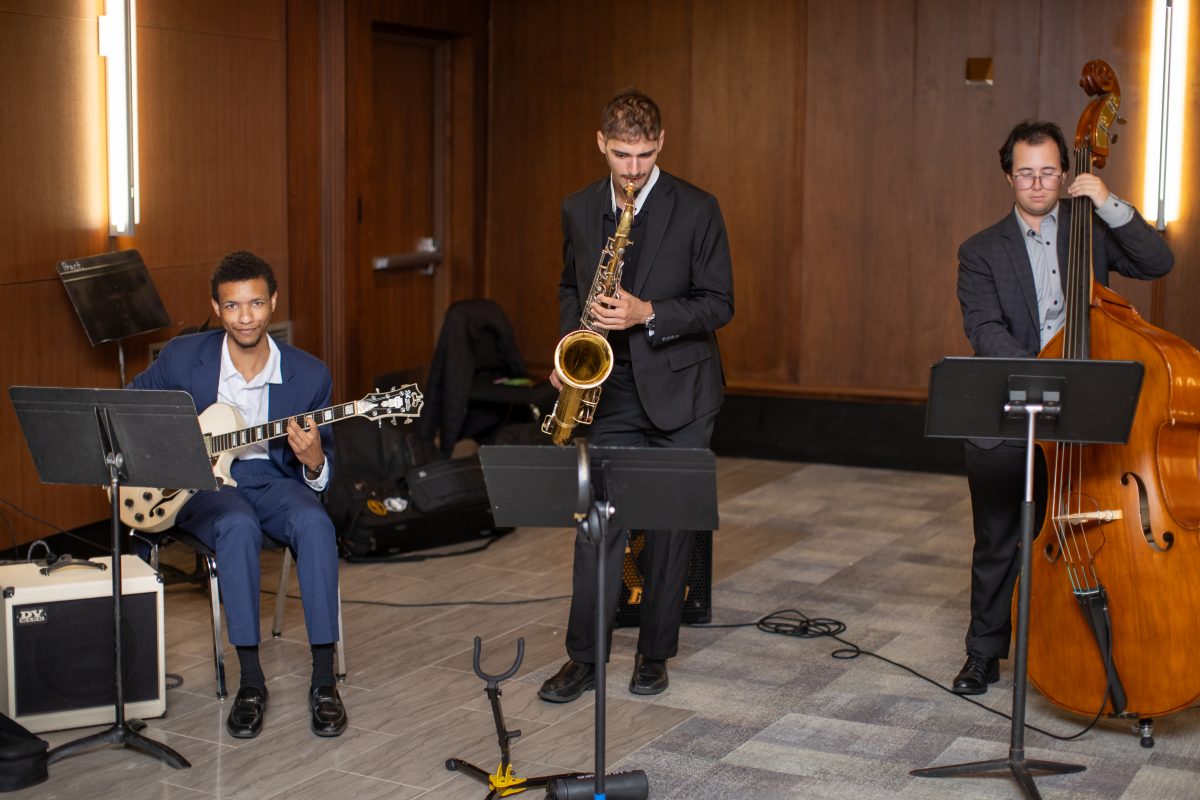 Three gentlemen playing music in a band. One is with a guitar, the other one is on the saxophone and the last one is on the cello.